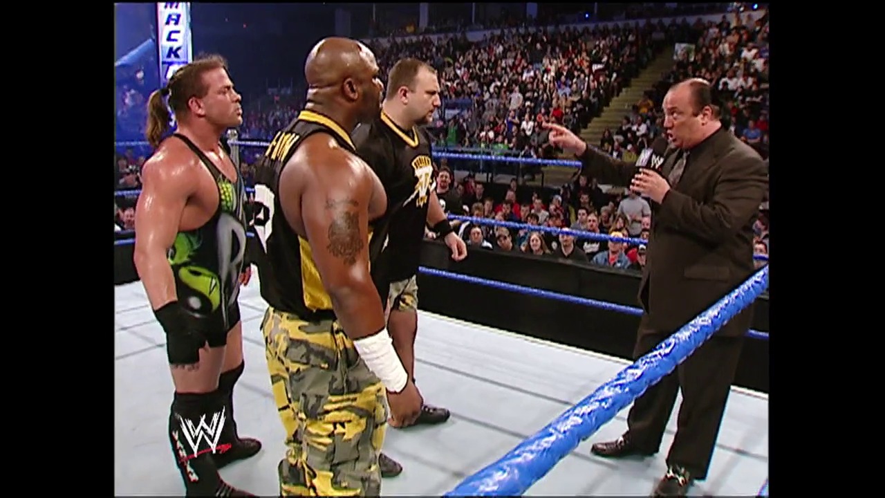 A frame from WWE SmackDown (April 22, 2004) at 13:11. A WWE SmackDown match scene with three wrestlers in the ring facing a man in a brown suit holding a microphone. The crowd fills the arena in the background, with blue ring ropes visible in the foreground.