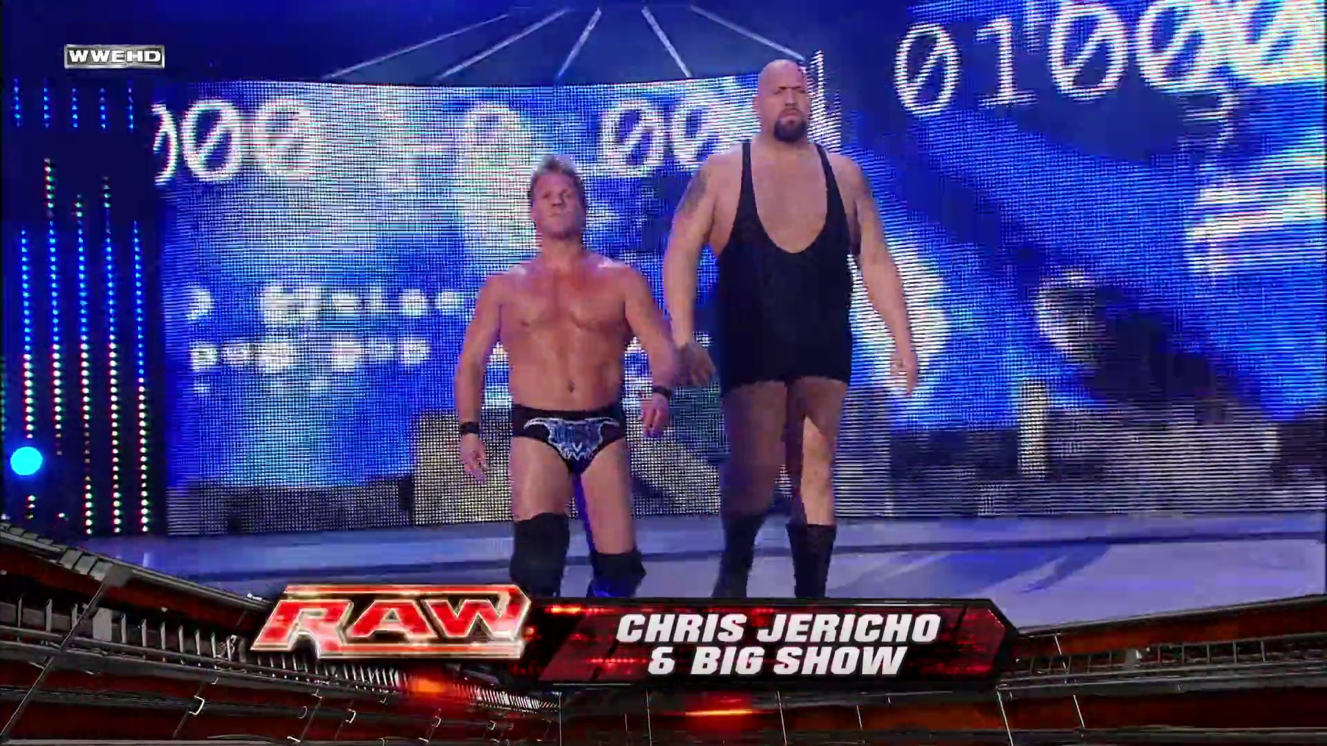 A frame from WWE Raw (January 4, 2010) at 40:33. Two wrestlers enter the ring during WWE Raw, with one shirtless in wrestling trunks and the other in black attire. The broadcast displays "CHRIS JERICHO 6 BIG SHOW" on the lower third, while blue LED screens and arena lighting frame the wrestling stage.