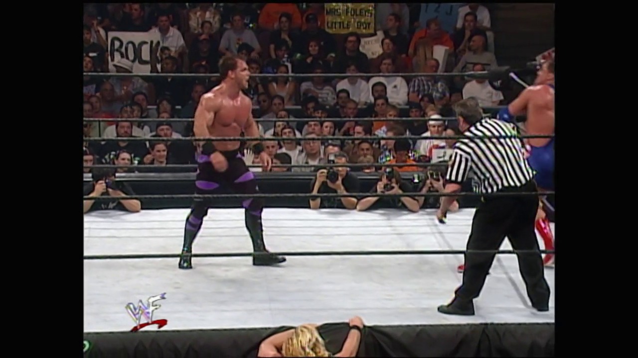 A frame from WrestleMania 2000 (2000) at 2:08:43. A professional wrestler in purple and black attire faces off against an opponent in the WWF ring, while a referee in striped shirt officiates. The crowd fills the arena background, with the WWF logo visible in the lower left corner of the broadcast.
