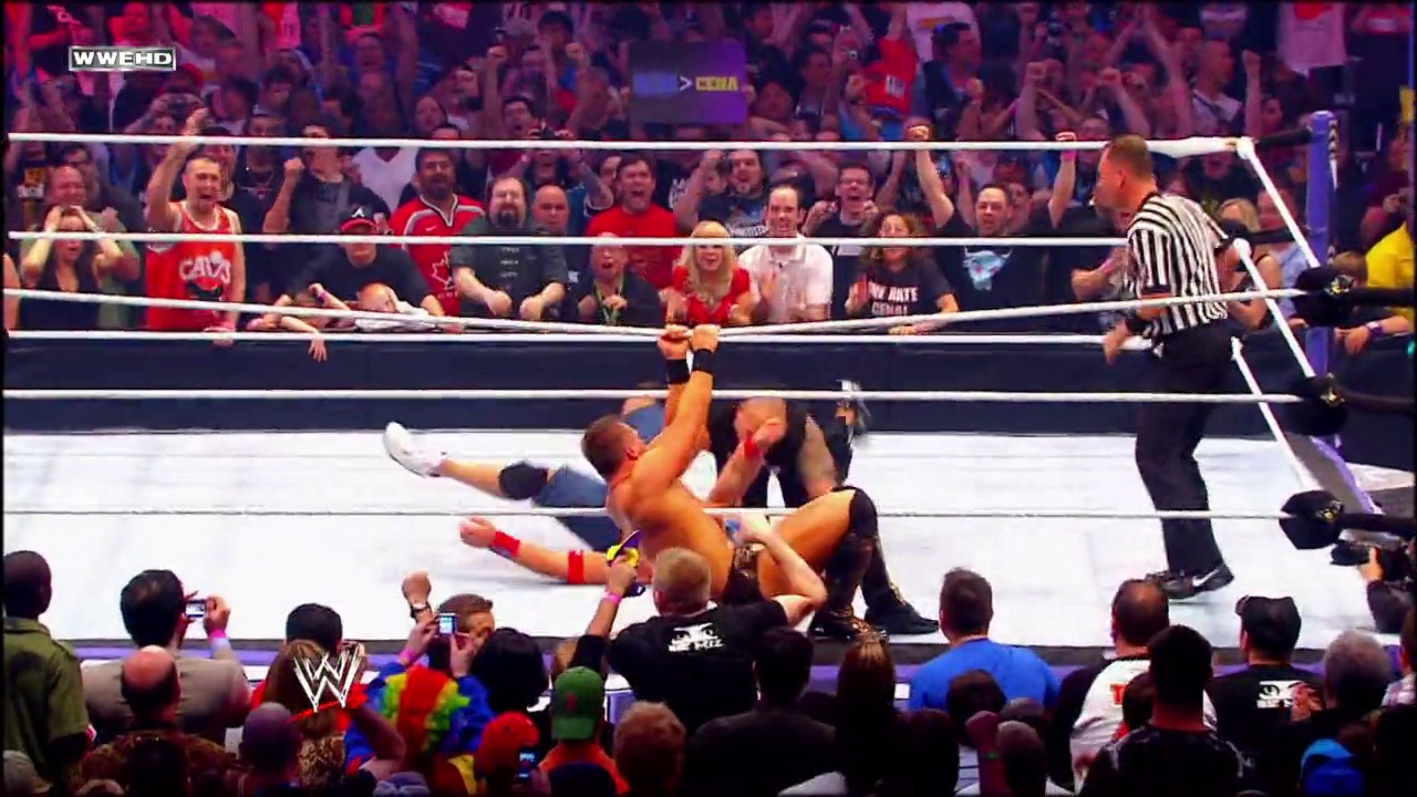 A frame from WrestleMania XXVI (2010) at 3:54:43. A WWE wrestler lies on the mat during an intense match at WrestleMania XXVI, with a referee nearby and an enthusiastic crowd visible behind the ring ropes. Fans capture the action on their phones while the packed arena creates an electric atmosphere.