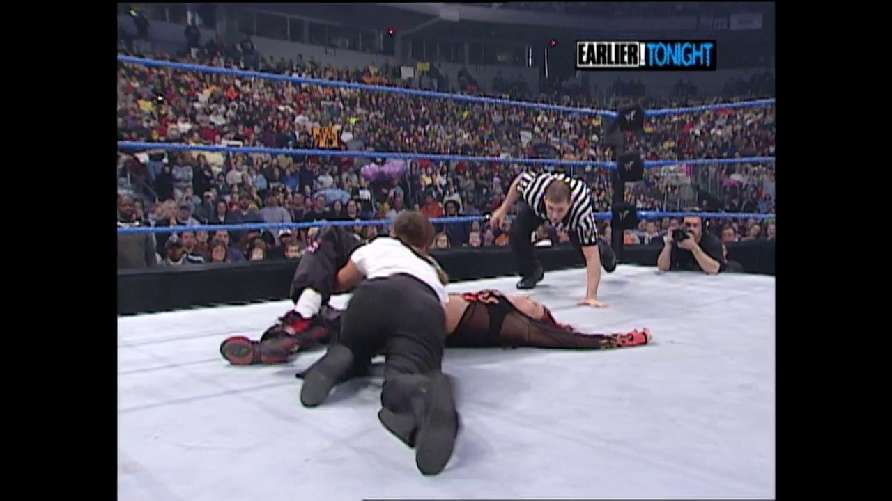 A frame from WWE SmackDown (December 14, 2000) at 1:03:08. A WWE wrestler in black and white attire pins an opponent in burgundy and black gear on the mat, while a referee in striped shirt oversees the match. The packed arena crowd fills the background under bright blue ring ropes, with an "EARLIER TONIGHT" graphic visible in the corner.