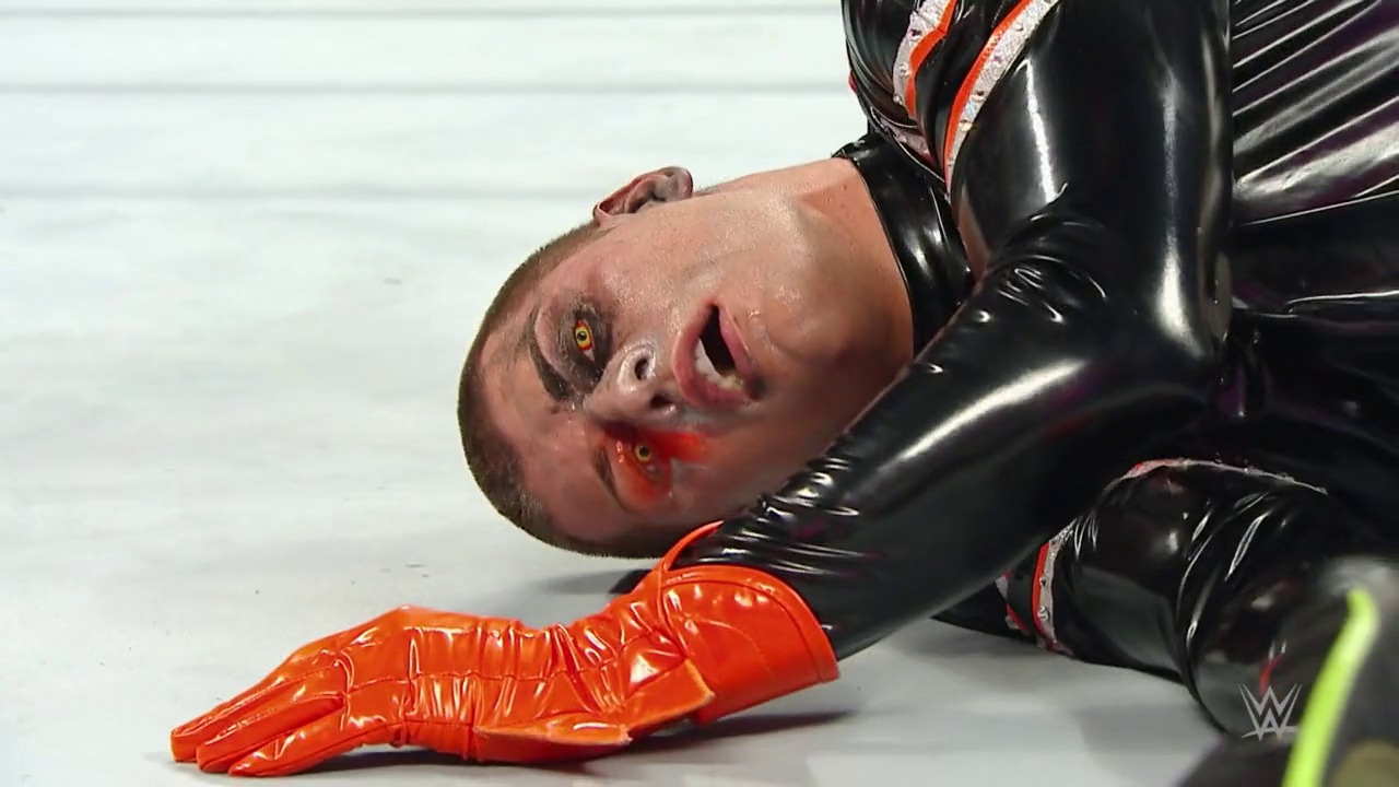 A frame from WWE Raw (April 6, 2015) at 56:14. A wrestler lies on the mat with visible facial injuries and blood, wearing black and orange wrestling attire. The WWE logo is visible in the corner, indicating this is from a professional wrestling broadcast.
