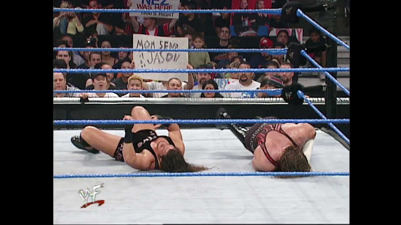 A frame from WWE SmackDown (May 24, 2001) at 1:04:11. Two wrestlers lie on the white canvas of a WWE SmackDown ring, surrounded by blue ropes. The crowd is visible in the background holding signs, with the WWF logo displayed in the bottom left corner.