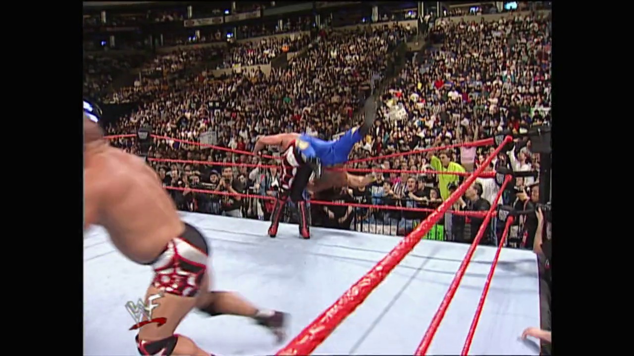 A frame from WrestleMania XIV (1998) at 13:15. A wrestler in blue performs an aerial move above the ring while another competitor lies on the mat below. The packed arena crowd watches intently from the stands surrounding the red-roped wrestling ring.