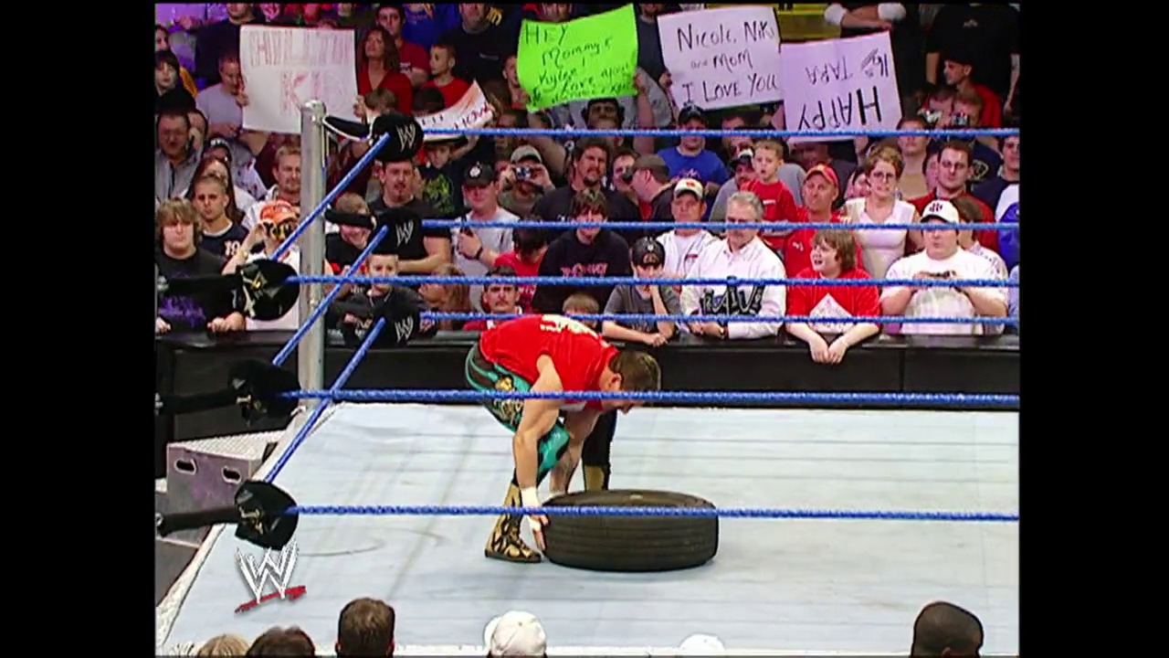 A frame from WWE SmackDown (December 9, 2004) at 24:10. A WWE wrestler in red and gold attire performs in the ring during SmackDown, with an engaged crowd holding signs visible in the background. The blue-roped wrestling ring, WWE graphics, and packed arena atmosphere capture the intensity of professional wrestling entertainment.