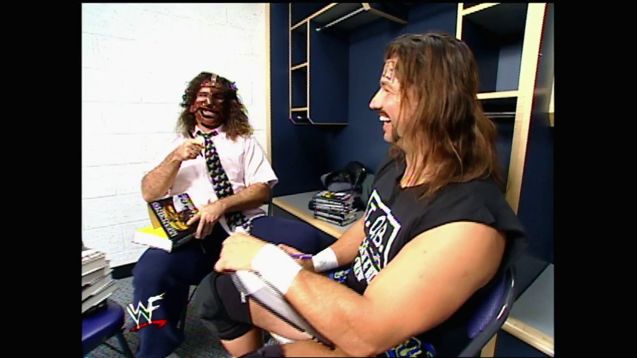 A frame from WWE SmackDown (December 9, 1999) at 35:45. Two wrestlers engage in a backstage interview in a WWF studio setting. One wears a white shirt with a decorative tie and face paint, while the other sits across from him in black attire, with stacked merchandise visible on a table between them.
