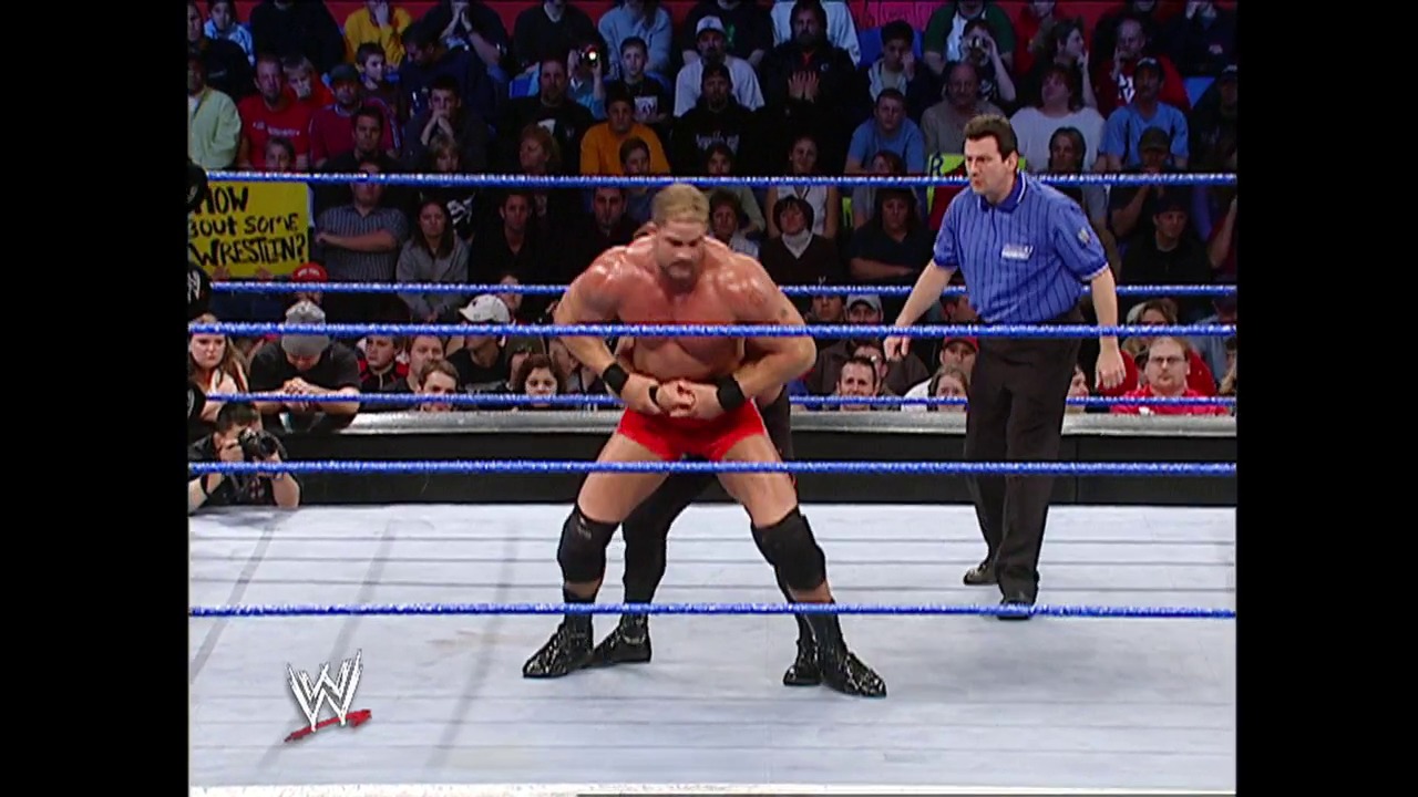 A frame from WWE SmackDown (November 27, 2003) at 12:19. A muscular wrestler in red shorts and black boots stands in a WWE wrestling ring, flexing in an aggressive stance. A referee in a blue shirt stands nearby as the crowd watches from the stands, with the WWE logo visible on the ring mat.