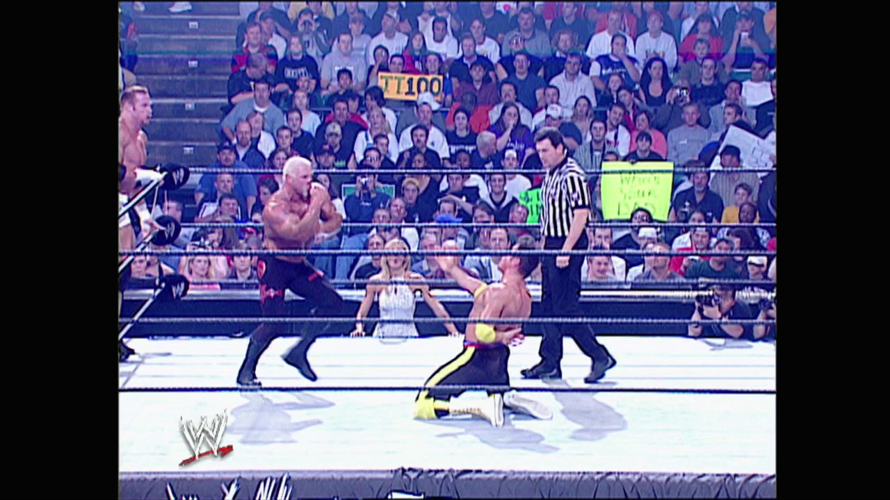 A frame from WWE Judgment Day (2003) at 23:40. A professional wrestler in black and yellow attire lies on the mat while a bald opponent stands nearby, with a referee in striped shirt overseeing the match. The packed arena crowd watches from the stands as the WWE logo appears in the corner.