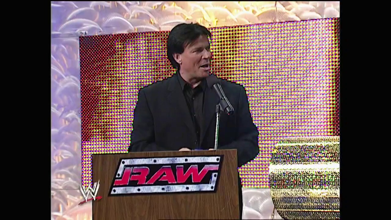 A frame from WWE Raw (March 22, 2004) at 42:05. A man in a dark suit stands at a wooden podium branded with the WWE Raw logo, speaking into a microphone against a colorful LED screen backdrop featuring red, yellow, and blue patterns.