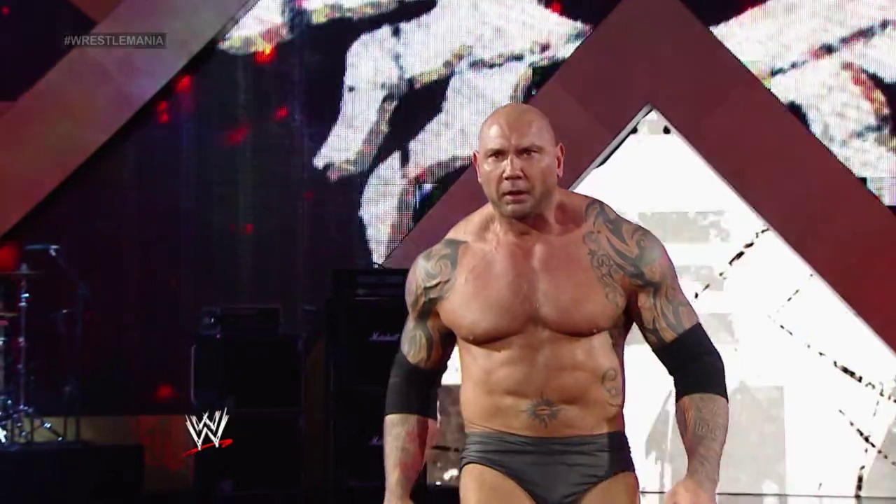 A frame from WrestleMania XXX (2014) at 3:19:44. A shirtless, tattooed wrestler stands in the ring during WrestleMania XXX, with the WWE logo visible in the lower left corner and large video screens displaying wrestling action in the background against a burgundy-toned stage setup.