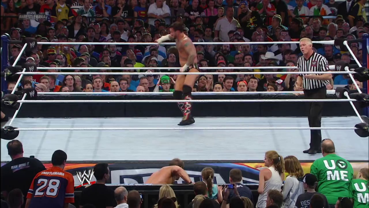 A frame from WrestleMania XXVIII (2012) at 2:44:56. A shirtless wrestler runs across the white WWE ring while a referee in black and white stripes watches. The crowd fills the arena around the ring, with visible signage including "#WrestleMania" and "WRESTLEMANIA 28" merchandise visible ringside.