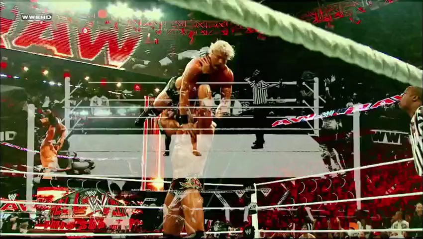 A frame from WWE Raw (November 7, 2011) at 26:07. A blonde wrestler performs a move from the top rope onto an opponent in the ring, while a referee watches from the side. The WWE Raw branding and red graphics are visible throughout the arena with spectators in the background.
