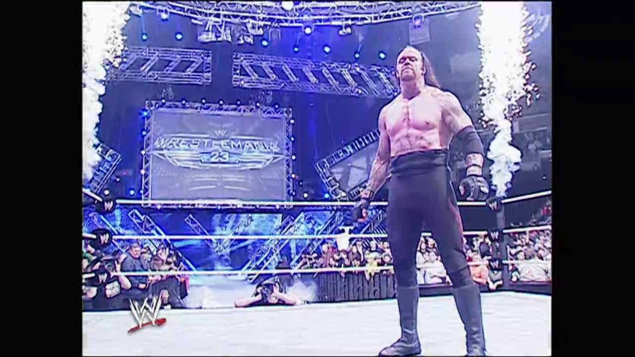 A frame from Royal Rumble (2007) at 2:43:41. A muscular wrestler stands in a WWE ring during WrestleMania 23, shirtless with arm bands, facing the crowd. The arena is illuminated with blue lighting and festive decorations, with packed bleachers visible in the background.