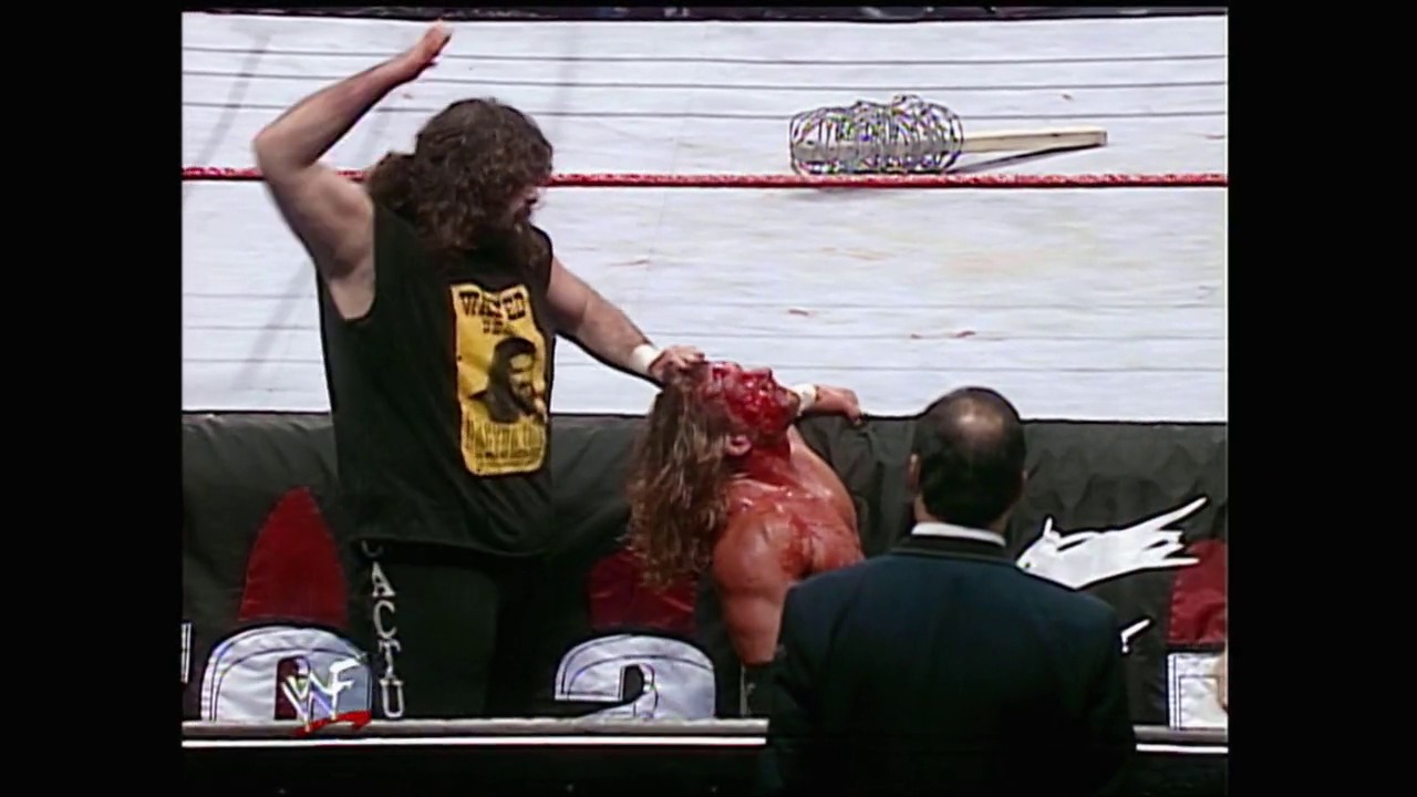 A frame from Royal Rumble (2000) at 1:23:20. A wrestler in a black and yellow shirt stands victorious in a WWF wrestling ring, with another competitor appearing bloodied below. Officials in dark suits are present ringside as the crowd watches the intense match action unfold.