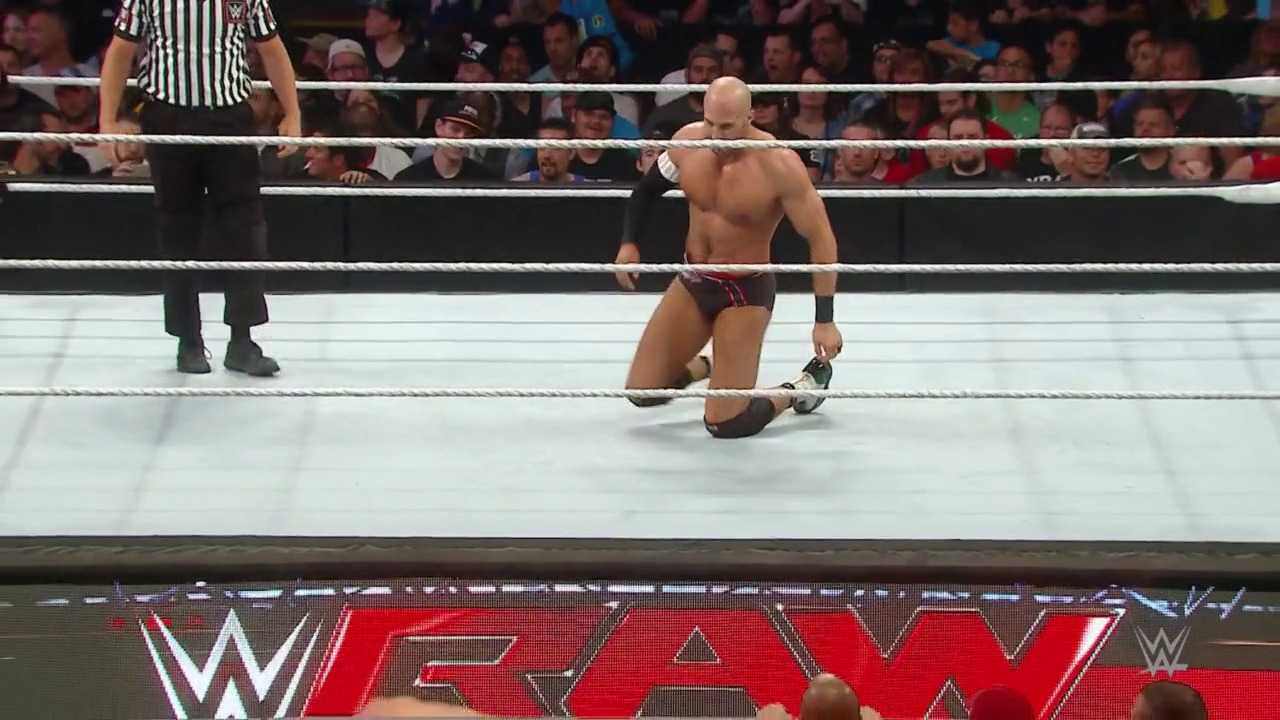 A frame from WWE Raw (July 6, 2015) at 2:02:52. A shirtless wrestler kneels in a WWE Raw ring surrounded by white ropes, with a referee in a striped shirt nearby. The crowd fills the background while the WWE Raw logo is visible on the digital signage below the ring.