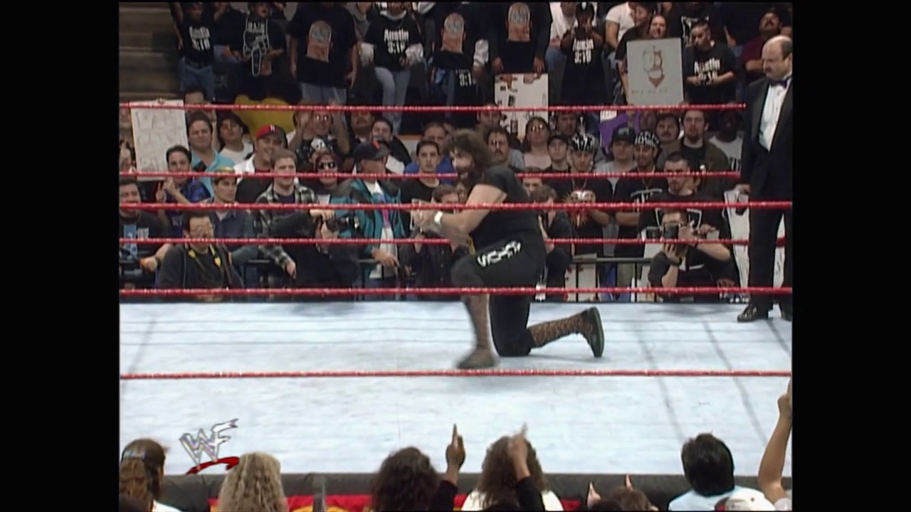 A frame from Royal Rumble (1998) at 1:10:57. A wrestler kneels in a WWE ring surrounded by red ropes, while enthusiastic fans cheer from the packed stands above. The crowd holds signs and wears event merchandise, with officials visible at ringside during this intense Royal Rumble moment.