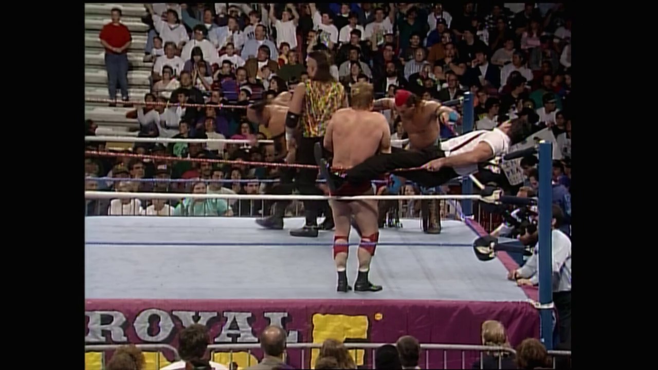 A frame from Royal Rumble (1993) at 2:06:09. A wrestler in a gold and black outfit stands in the ring while another competitor is being thrown over the top rope during a Royal Rumble match, with a packed crowd visible in the bleachers behind the burgundy-padded ring apron.