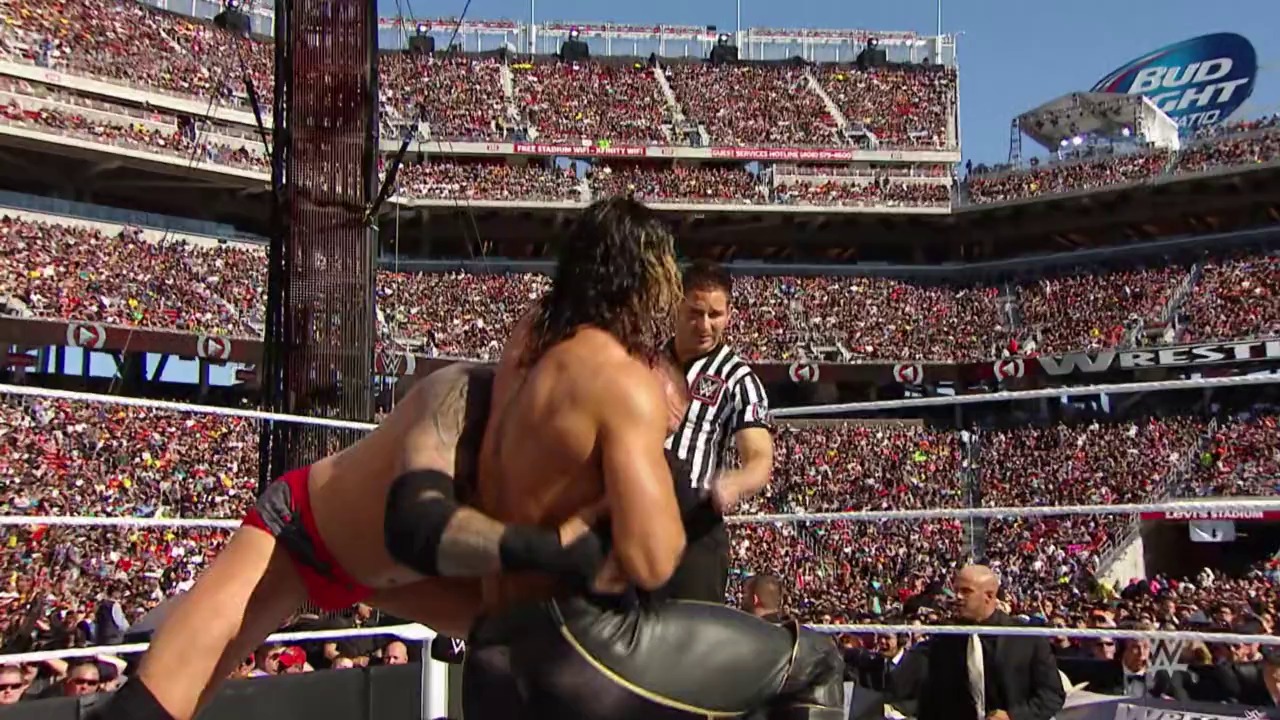 A frame from WrestleMania (2015) at 35:06. A wrestler in red and black attire executes a move against an opponent in the ring during WrestleMania, with a referee present and a massive crowd filling the stadium under clear skies. The Bud Light blimp is visible above the packed venue.