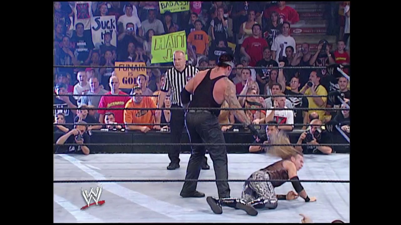 A frame from Summerslam (2002) at 1:32:59. A WWE wrestler stands in the ring while another lies on the mat, with a referee in striped shirt officiating. The crowd fills the arena background, displaying signs including one reading "MAIN," as the WWE logo appears in the lower left corner.