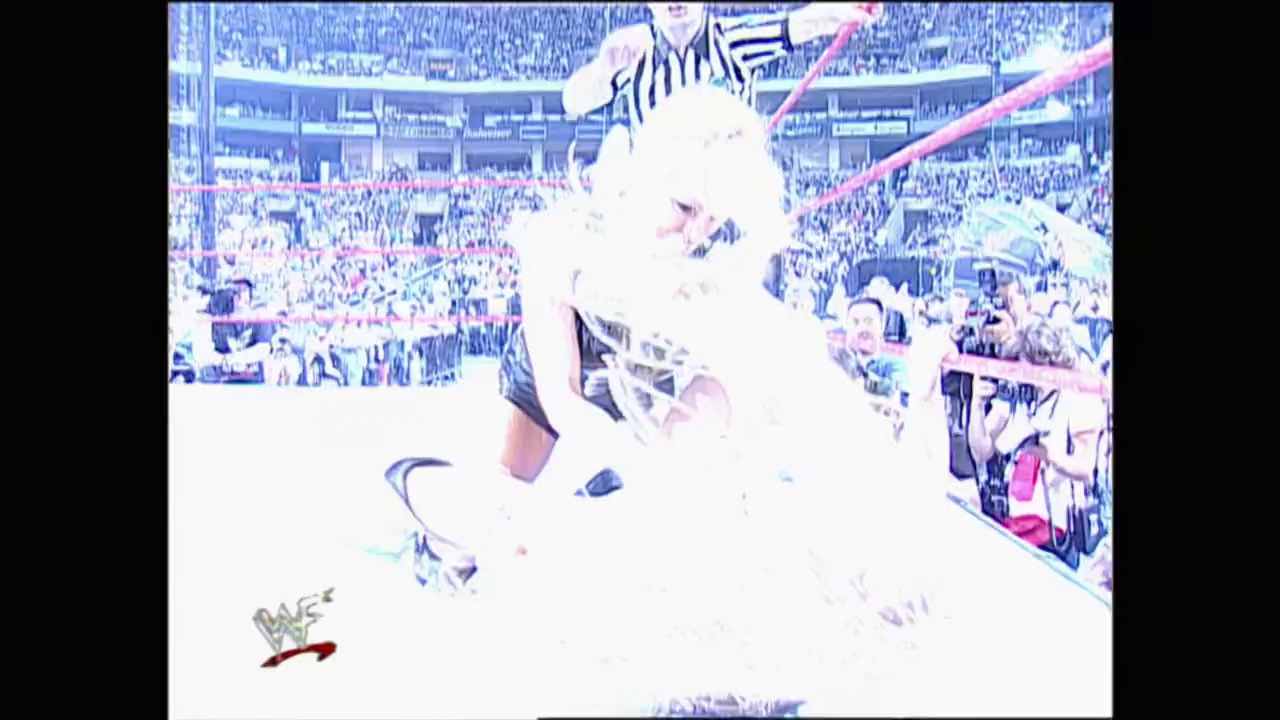A frame from WrestleMania XIV (1998) at 54:05. A wrestler performs a high-flying move above the ring at WrestleMania XIV, with a packed stadium crowd visible in the background and on-screen graphics displayed around the arena.