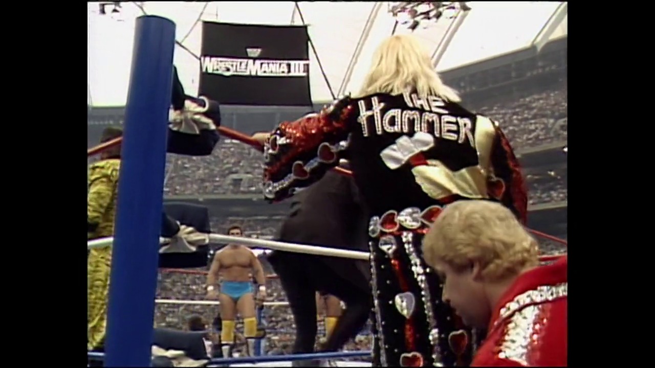 A frame from WrestleMania III (1987) at 53:28. A wrestler in an ornate red and black outfit with "The Hammer" on the back stands in a WrestleMania III ring, facing an opponent in blue trunks, with a packed stadium crowd visible in the background.