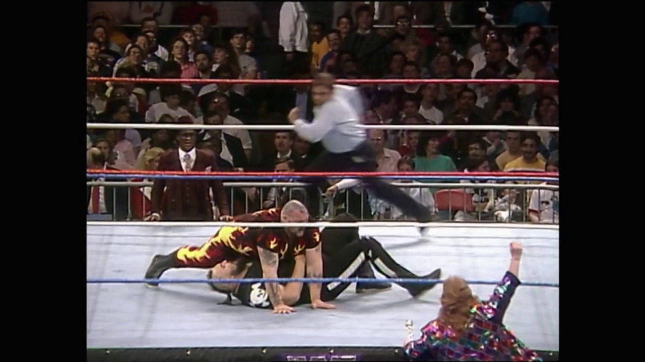 A frame from WrestleMania IV (1988) at 1:06:42. A wrestler in a yellow and red outfit lies on the mat after being thrown over the ropes, while an opponent in colorful attire celebrates below. The packed arena crowd watches from the stands as referees monitor the action in the WWF ring.