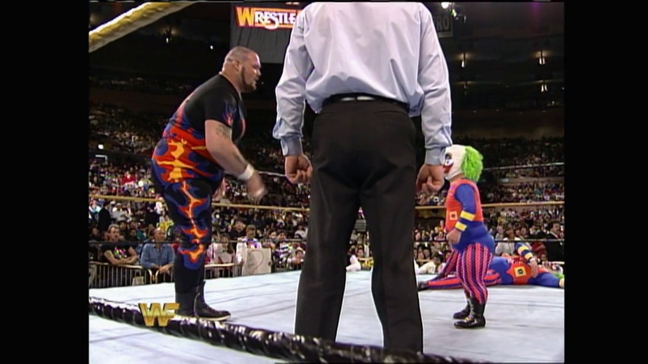 A frame from WrestleMania X (1994) at 46:45. A WWE wrestler in a colorful flame-patterned outfit stands in the ring facing a referee in a white shirt, while a smaller performer in a clown costume sits on the mat nearby. The packed arena crowd is visible in the background with WrestleMania signage overhead.