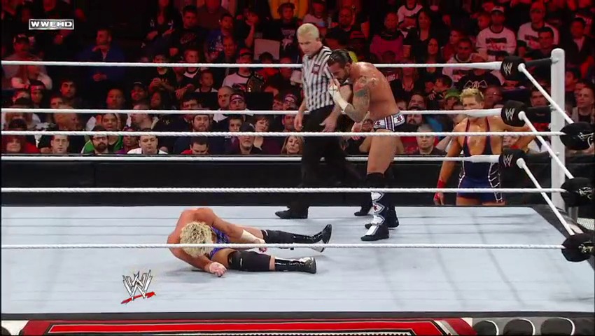 A frame from WWE Raw (December 26, 2011) at 1:21:36. A WWE wrestler lies on the mat after being knocked down, while their opponent stands nearby. A referee in a striped shirt oversees the match as the crowd watches from the stands in the red-lit arena.