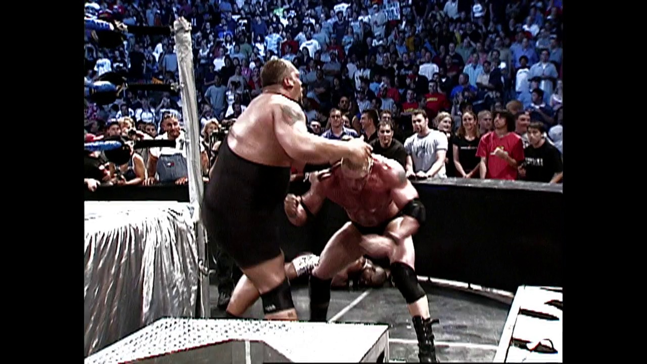 A frame from WWE SmackDown (July 17, 2003) at 0:52. Two wrestlers grapple in a WWE ring during an intense match, with one performing a move against his opponent near the corner post. A large crowd of enthusiastic fans fills the arena stands in the background.