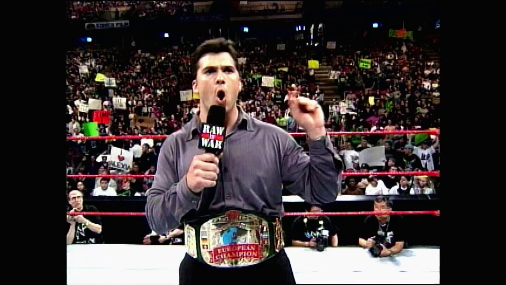 A frame from WWE Raw (April 19, 1999) at 0:47. A wrestler stands in the WWE Raw ring holding a microphone, wearing a dark shirt and an European Championship belt, addressing a packed arena crowd with an animated expression. The red-roped ring and enthusiastic fans with signs fill the background.