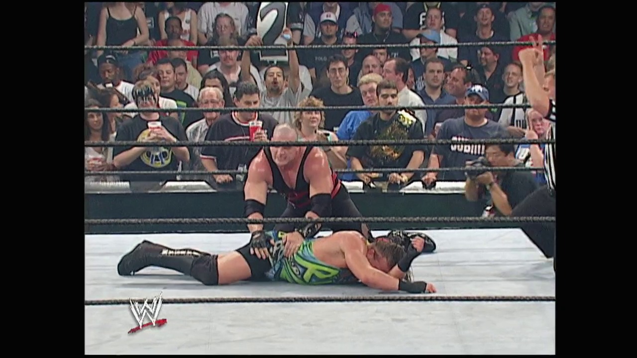 A frame from Summerslam (2003) at 1:57:24. A WWE wrestler in black and neon green attire lies on the mat after being thrown, while an opponent stands nearby. The crowd watches intently from the stands surrounding the white ring, with the WWE logo visible at the bottom of the frame.