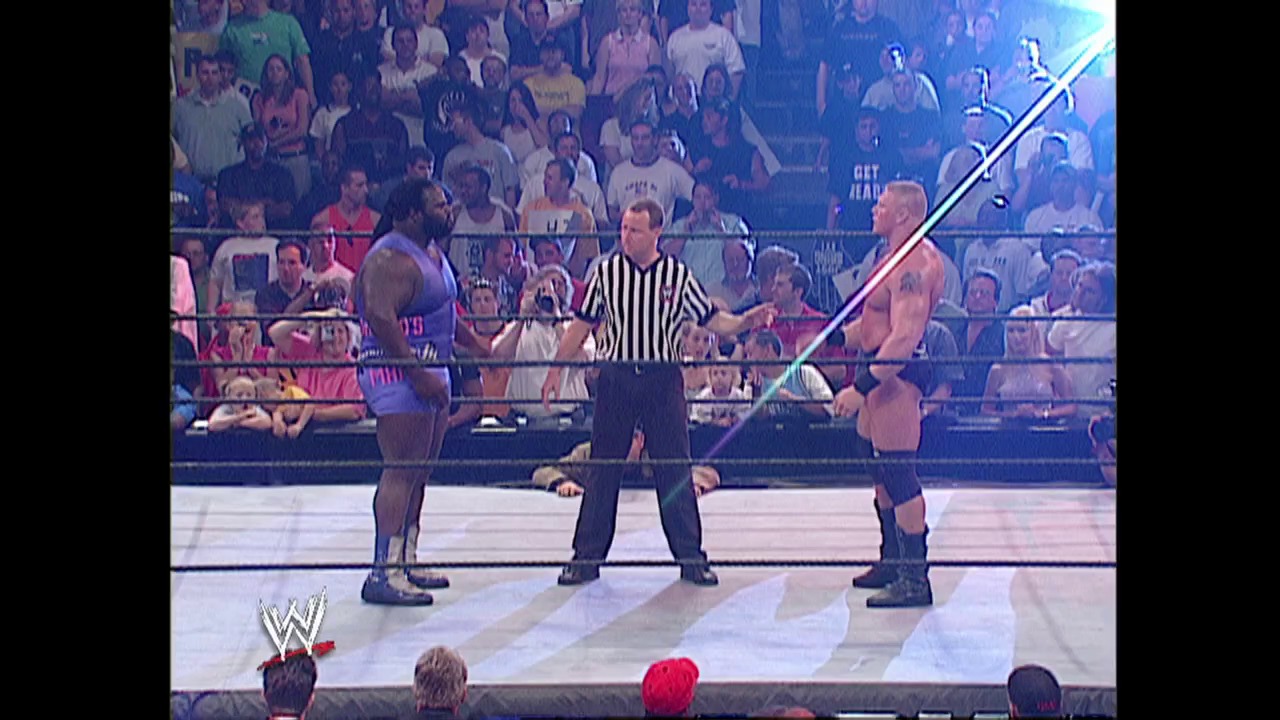 A frame from WWE Raw (August 1, 2002) at 25:10. A WWE wrestling match is in progress with two shirtless wrestlers facing off in the ring while a referee in a striped shirt stands between them. The crowd fills the arena in the background under blue lighting, with the WWE logo visible in the lower left corner.