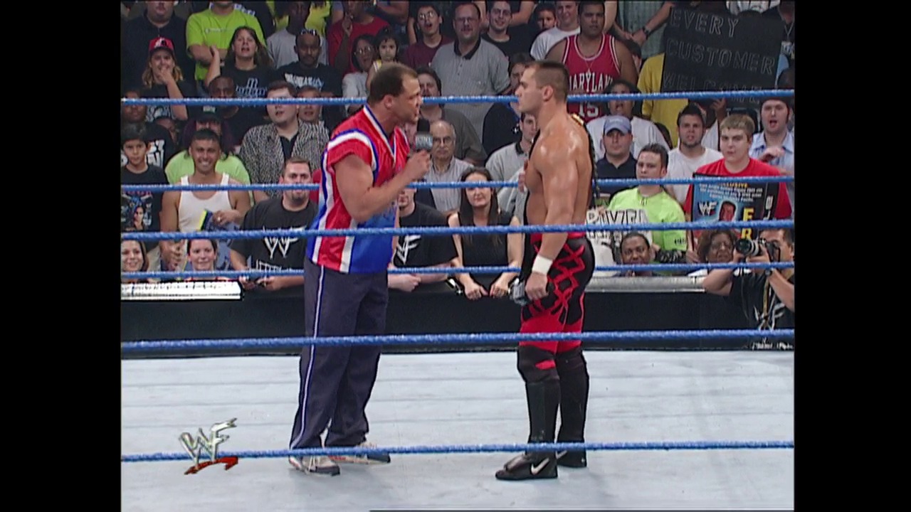 A frame from WWE SmackDown (August 2, 2001) at 13:02. A referee in a red and white shirt conducts an interview with a shirtless wrestler wearing red and black pants in a WWE SmackDown ring, surrounded by enthusiastic fans and blue ropes.