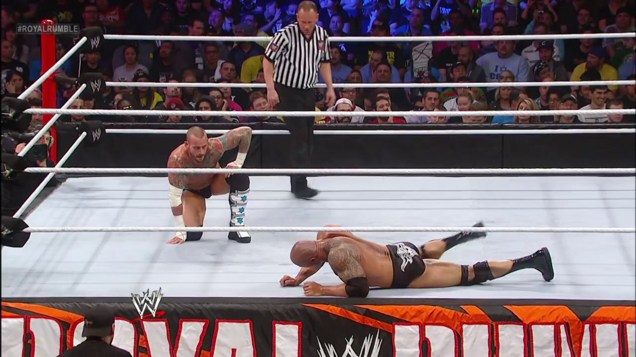 A frame from Royal Rumble (2013) at 2:22:55. A WWE wrestler lies on the mat while another approaches during a Royal Rumble match, with a referee overseeing the action in the brightly lit ring surrounded by an enthusiastic crowd.