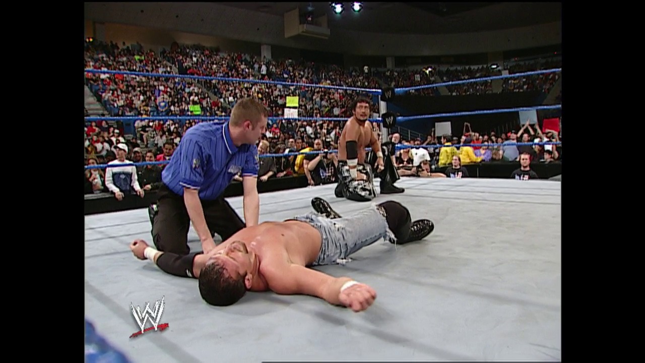 A frame from WWE SmackDown (January 9, 2003) at 37:13. # WWE SmackDown Ring Action

A referee in blue officiates as a shirtless wrestler lies prone on the mat while another competitor stands nearby, with a packed arena crowd visible in the background and the WWE logo displayed in the corner.
