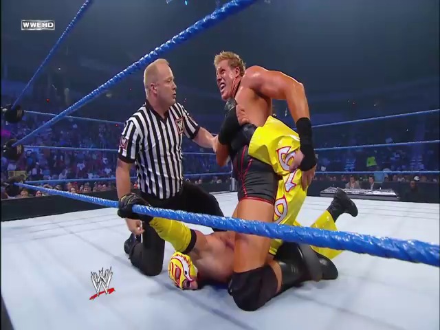 A frame from WWE SmackDown (July 23, 2010) at 1:12:41. A WWE wrestler in yellow and black attire is pinned to the mat by an opponent in a black singlet, while the referee in a striped shirt makes the count during a SmackDown match, with the blue-roped ring and crowd visible in the background.