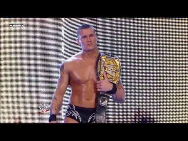 A frame from WWE Raw (March 3, 2008) at 1:15:27. A shirtless WWE wrestler displays a championship belt on his shoulder while standing on a large LED screen during a televised event. The performer is muscular and posed confidently against the bright digital backdrop.
