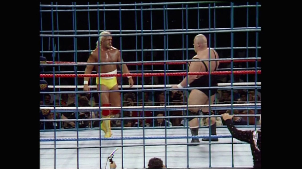 A frame from WrestleMania 2 (1986) at 2:48:19. Two wrestlers face off in a steel cage match inside a WWE ring, with red ropes visible and a crowd watching from below. The wrestler on the left wears yellow and black attire with a headband, while his opponent on the right is shirtless.