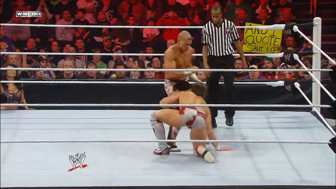 A frame from WWE Raw (January 31, 2011) at 38:36. A WWE wrestler in red and white attire performs a wrestling move in the ring while an opponent stands nearby. A referee in striped shirt supervises as the crowd watches from the stands, with the WWE logo visible on the mat.