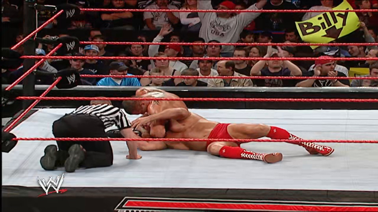A frame from WWE Raw (November 26, 2007) at 1:25:24. A WWE wrestler in red attire lies on the mat while a referee in striped shirt tends to them during a Raw event. The crowd watches from the ringside as the match unfolds in the iconic red-roped WWE ring.