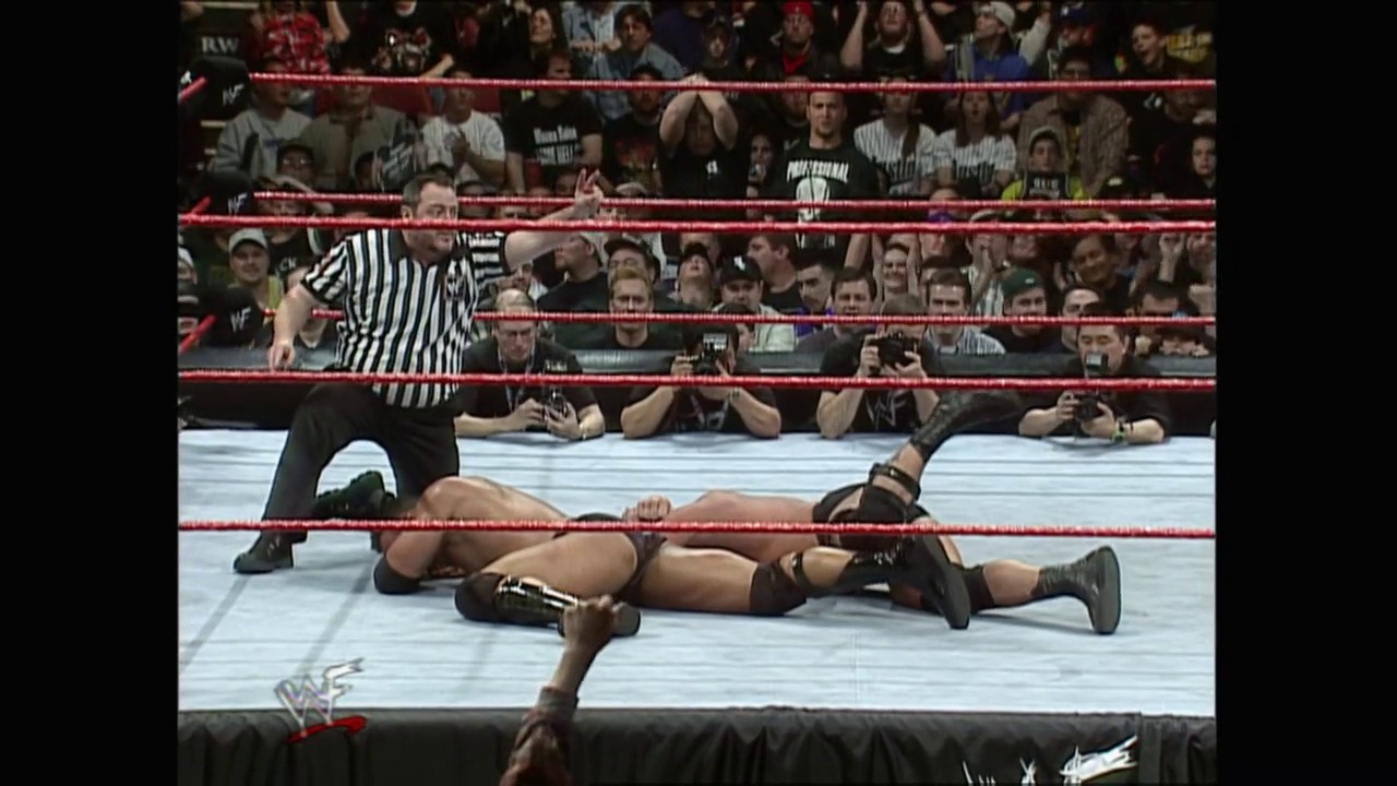 A frame from WrestleMania XV (1999) at 2:32:34. A wrestler lies on the mat while a referee in striped shirt stands nearby during a WWF match. The red-roped ring is surrounded by an engaged crowd, with the distinctive WWF logo visible on the ring apron.