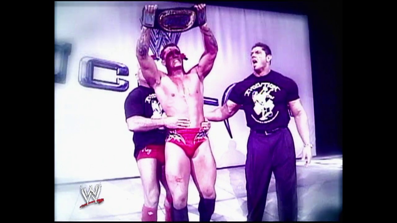 A frame from WWE Raw (May 10, 2004) at 13:52. A shirtless wrestler holds a championship belt above his head while two other wrestlers in black shirts stand beside him in the WWE ring. The WWE logo is visible in the bottom left corner of the broadcast.