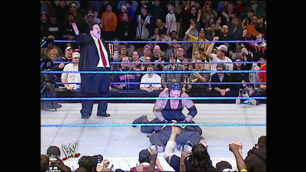 A frame from WWE SmackDown (March 18, 2004) at 1:28:18. A WWE wrestler in pink and blue attire sits on the canvas while another performer stands nearby in a suit. The blue-roped ring is surrounded by a large crowd of spectators, with the WWE logo visible in the lower left corner.
