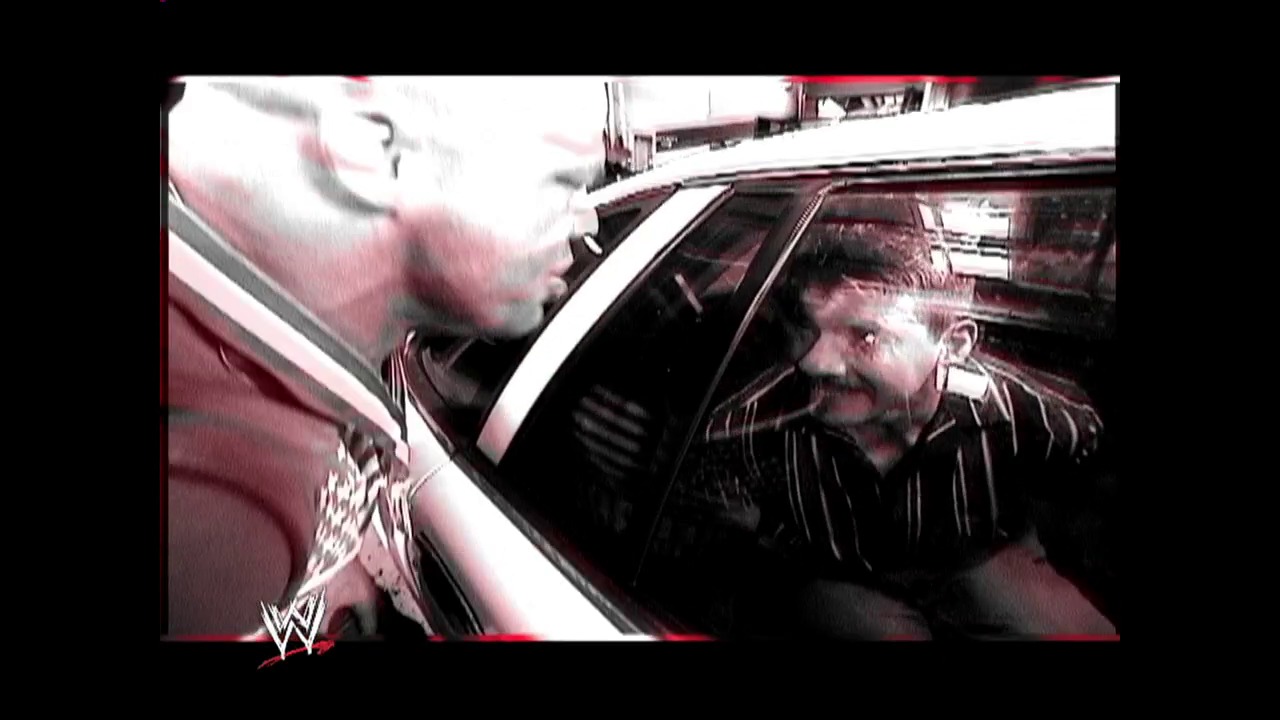 A frame from WWE SmackDown (March 4, 2004) at 0:57. A WWE SmackDown scene captures two men in conversation beside a car, with one wearing a pinstriped shirt. The WWE logo appears in the lower left corner of the broadcast frame.