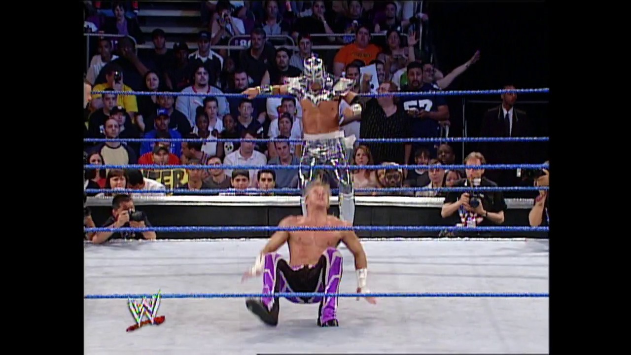 A frame from WWE SmackDown (June 26, 2003) at 1:01:00. A WWE wrestler in purple and black attire performs in the ring during SmackDown, with the crowd visible in the bleachers behind the blue-roped ring and WWE graphics displayed in the lower left corner.