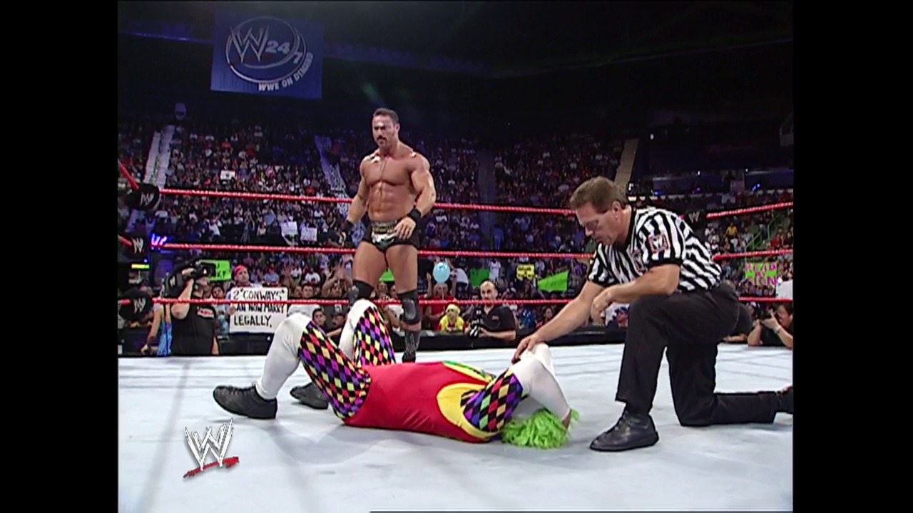 A frame from WWE Raw (October 10, 2005) at 14:00. A WWE wrestler in colorful patterned attire lies on the mat while a referee checks on him, with an opponent standing nearby. The crowd fills the arena in the background, and the red WWE ring ropes are visible in this professional wrestling match scene.