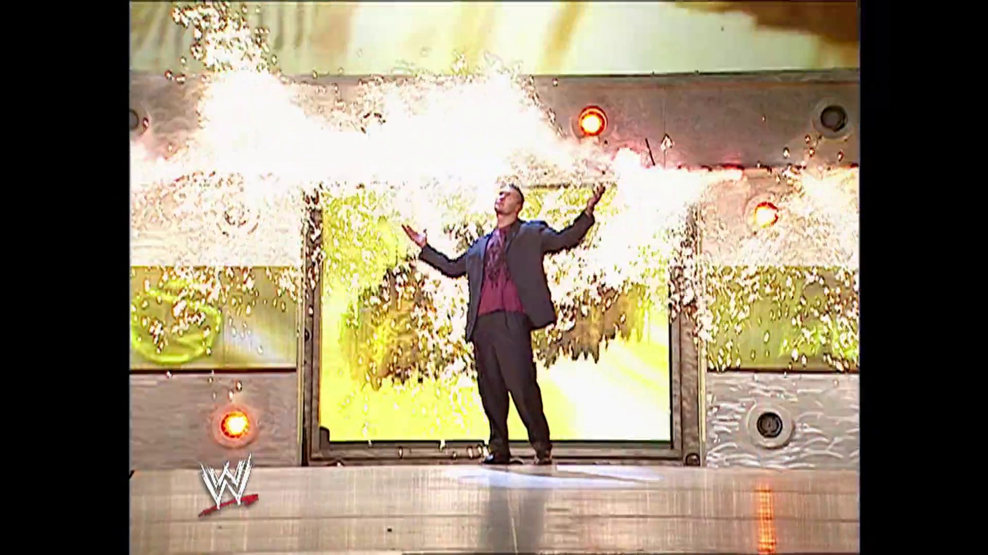 A frame from WWE Raw (October 9, 2006) at 1:51:17. A WWE performer stands in the center of the ring with arms raised, surrounded by explosive pyrotechnics and bright yellow flames. The stage features large video screens displaying nature imagery, with the WWE logo visible in the lower left corner.