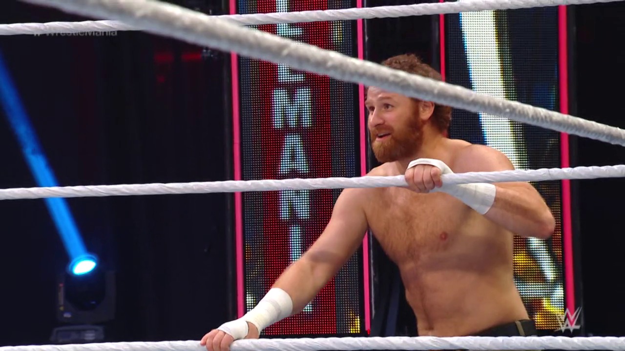 A frame from WrestleMania 36 (2020) at 1:10:52. A shirtless wrestler with reddish-brown hair and beard stands in a WWE ring, leaning against the ropes with white wrist tape. Colorful LED graphics and lighting are visible in the background behind the ring.