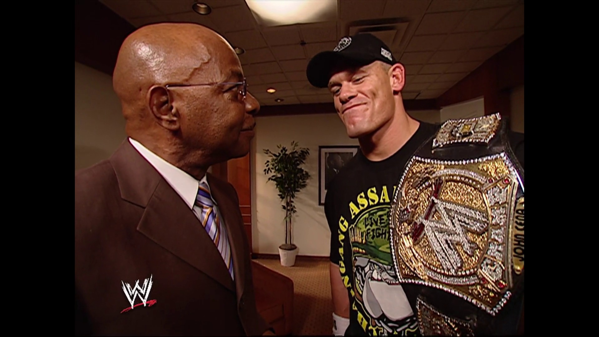 A frame from WWE SmackDown (September 22, 2006) at 1:05:41. A WWE backstage scene featuring two men in conversation: one wearing a brown suit and glasses on the left, and a wrestler on the right holding an ornate championship belt, wearing a black shirt and cap. The WWE logo is visible in the lower left corner.