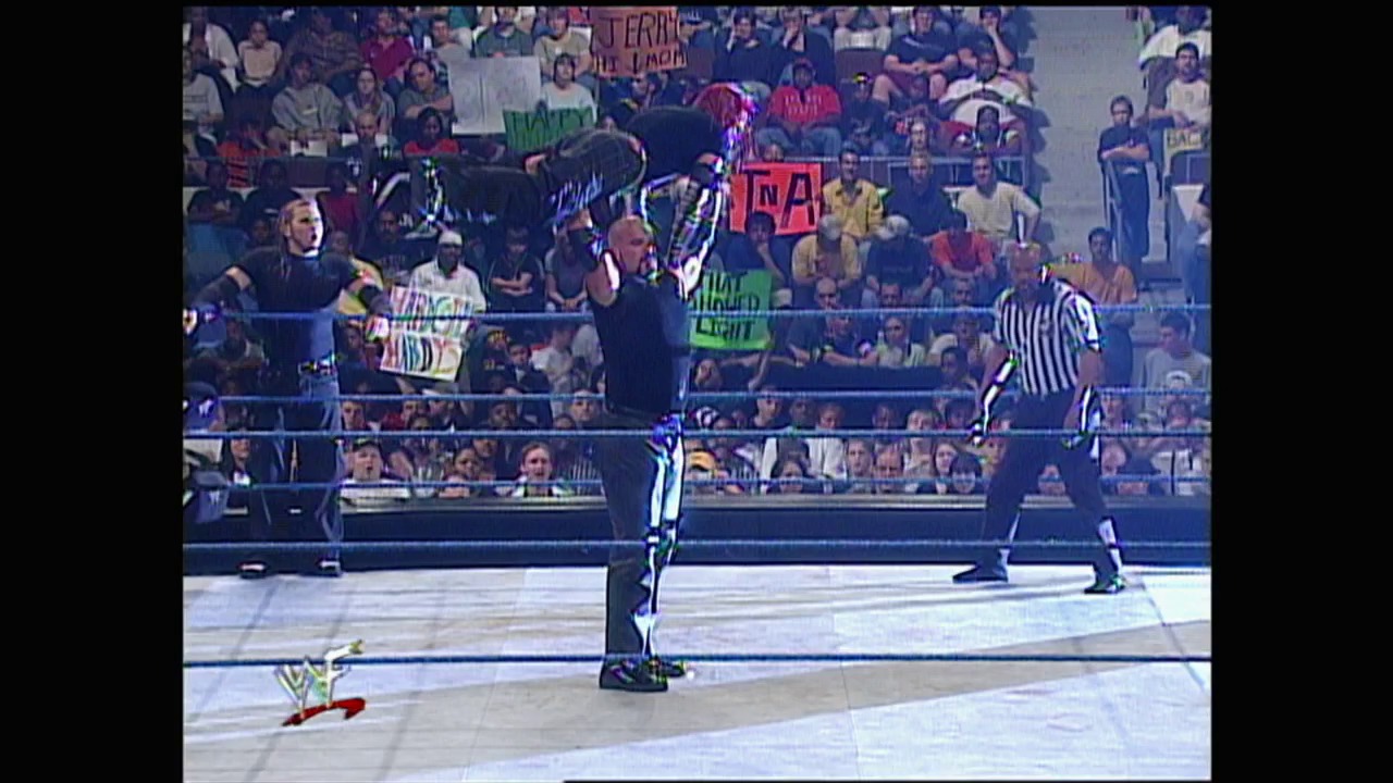 A frame from WWE SmackDown (May 4, 2000) at 49:09. A bald wrestler in dark attire stands in the WWE ring holding championship belts above his head, while a referee in striped shirt observes. The crowd fills the arena stands in the background with various signs visible.